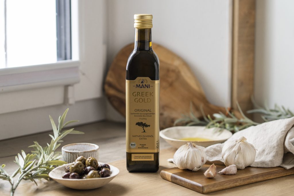 Greek Gold Original Olivenöl in mediterranem Setting mit Olivenzweig, Schälchen Oliven, Öl und Knoblauch