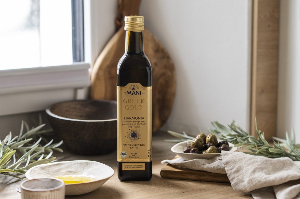 Greek Gold Olivenöl Harmonia in mediterranem Setting mit Olivenzweig, Schälchen Oliven und Öl